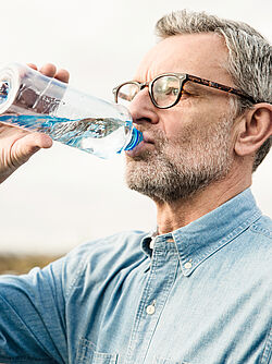 A man wearing glasses and a blue shirt drinks water from a bottle.