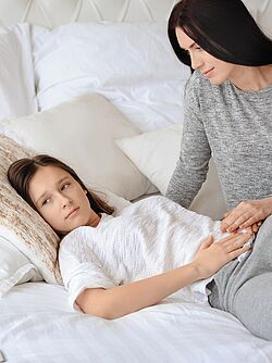 Teenage girl lying on a bed looking unwell, with her mother sitting beside her, providing comfort.