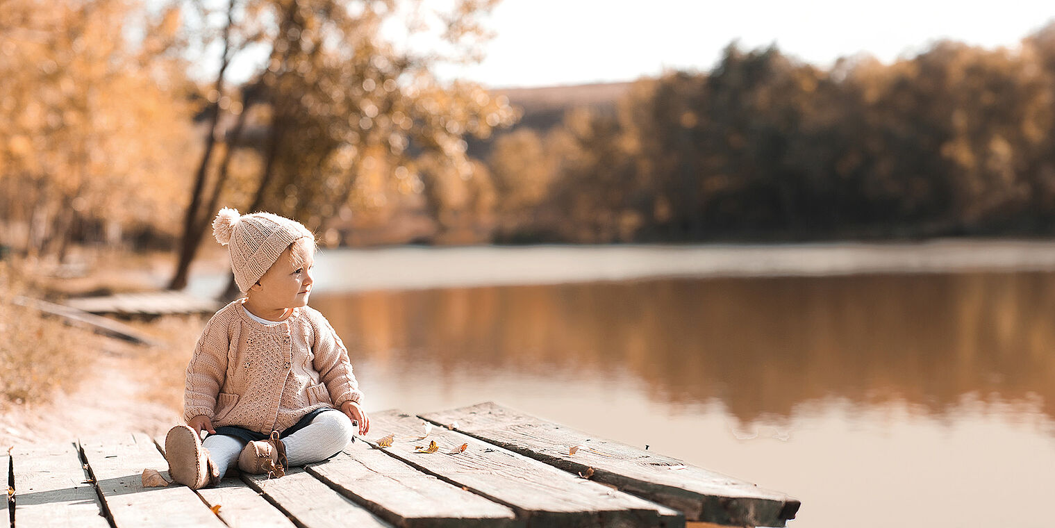 Baby is wearing a knitted hat with a pom-pom and sitting on a wooden dock by a calm lake